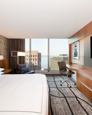 This image shows a modern hotel room with a large bed, a desk, a flat-screen TV, and a floor-to-ceiling window offering a city view.