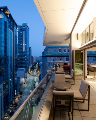 The image shows a modern rooftop terrace with seating and tables, overlooking a cityscape with tall buildings at dusk.