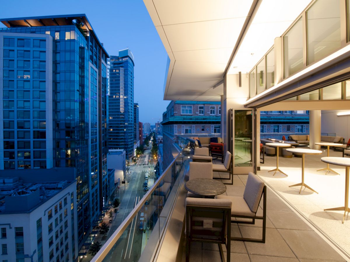 This image features a modern cityscape view from an elevated balcony with seating arrangements and tables, taken in the early evening.
