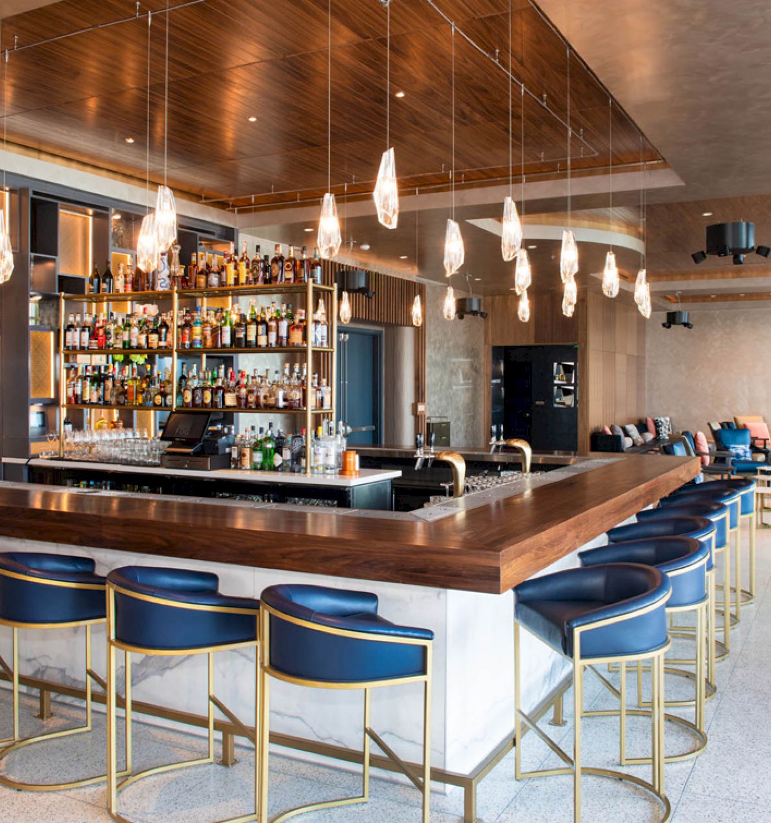 The image shows a modern bar with blue upholstered bar stools, a well-stocked liquor shelf, hanging lights, and a spacious seating area in the background.