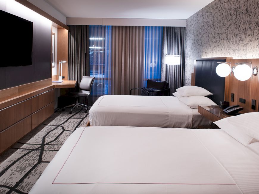 The image shows a modern hotel room with two neatly made beds, a wall-mounted TV, a work desk with a chair, and large windows with curtains.