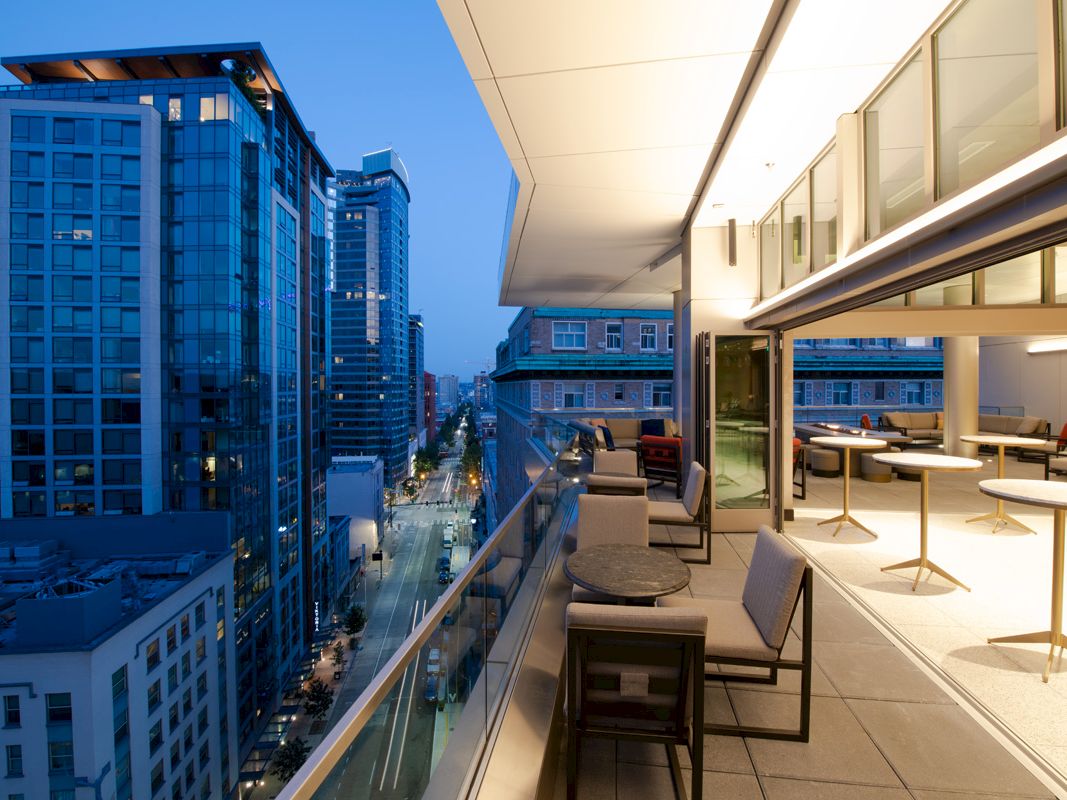 The image shows a modern balcony with seating and tables overlooking a cityscape featuring tall buildings and illuminated streets at dusk.