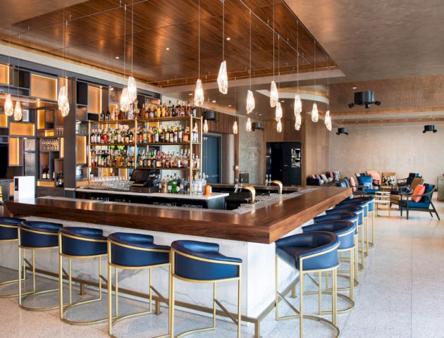 The image shows a modern bar with a well-stocked counter, surrounded by blue bar stools and featuring hanging pendant lights, and a seating area nearby.