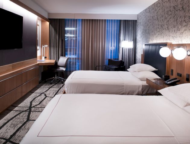A modern hotel room features two neatly made beds, a wall-mounted TV, a work desk with a chair, lamps, and a patterned carpet, with large windows.