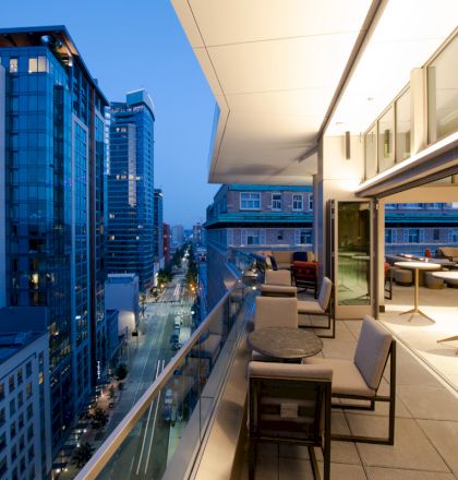 The image shows a modern balcony with outdoor seating overlooking a cityscape with high-rise buildings at dusk.