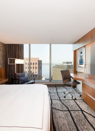 A modern hotel room with a large bed, a TV, a desk and chair, and a view of city buildings through a large window.