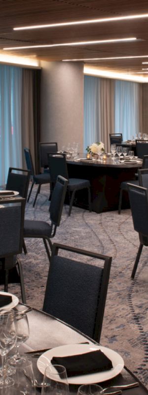 A modern banquet hall with round tables set with cutlery, glasses, and napkins, decorated with floral centerpieces and candles, and featuring blue curtains.