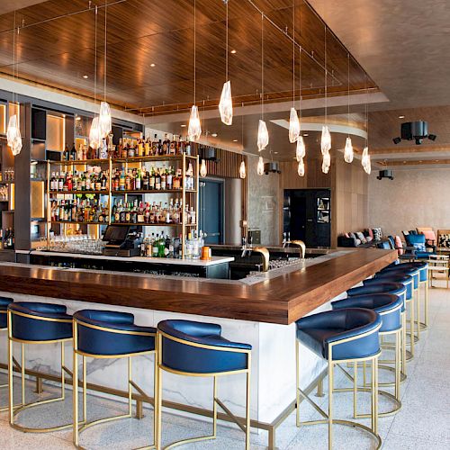 A modern bar with a wooden counter, blue bar stools, a well-stocked shelf of liquor, and stylish pendant lights. The interior is sleek and inviting.