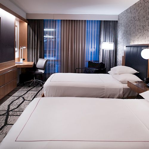 The image shows a modern hotel room with two double beds, a desk with a chair, a flat-screen TV, and a window with curtains.