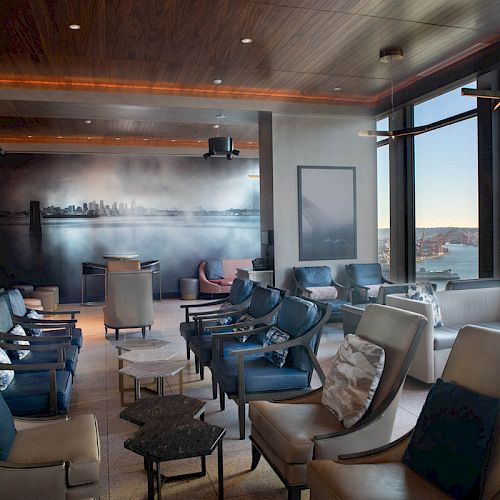 This image shows a modern lounge area with comfortable seating, a wall mural, large windows overlooking a waterfront view, and a Ferris wheel.