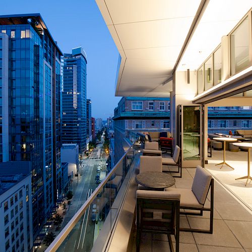 A modern city skyline at dusk with tall buildings. An outdoor terrace with tables and chairs, overlooking a lit street below.