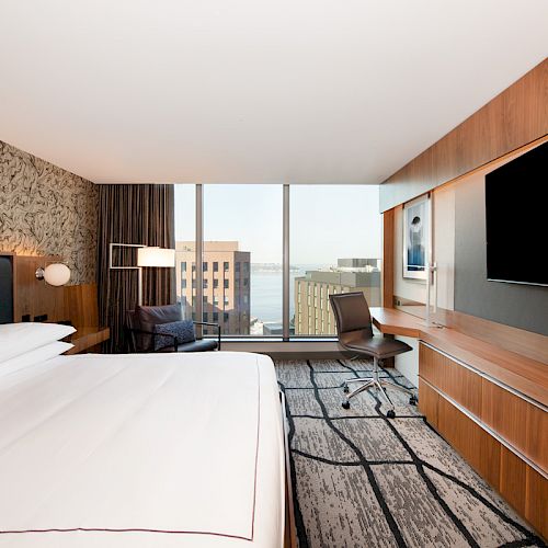 A modern hotel room features a large bed, a wall-mounted TV, a desk with a chair, and a window with a city view in the background.