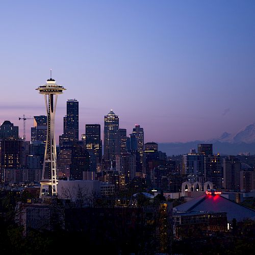 A cityscape at dusk featuring a prominent tower, with a mountain in the background and a mix of modern buildings in the foreground.