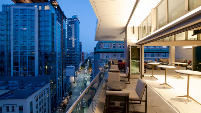 A modern cityscape at dusk featuring a high-rise balcony with seating and tables, overlooking a street lined with illuminated buildings.