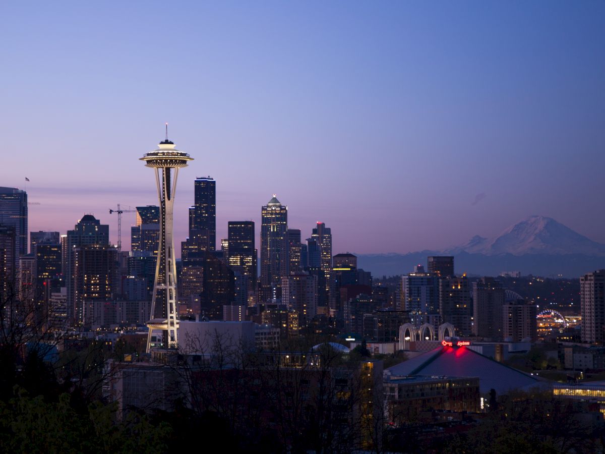 A twilight cityscape featuring the iconic Space Needle, surrounded by tall buildings. In the background, a mountain is visible against the purple sky.