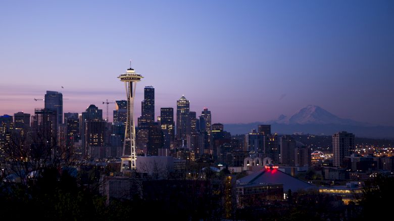 A twilight cityscape featuring the iconic Space Needle, surrounded by tall buildings. In the background, a mountain is visible against the purple sky.