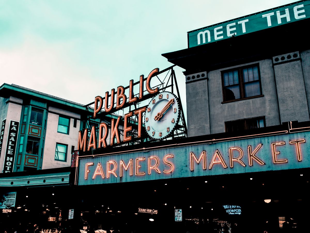 The image shows a public farmers market with neon signs, an iconic clock, and buildings with signs in the background under a cloudy sky.