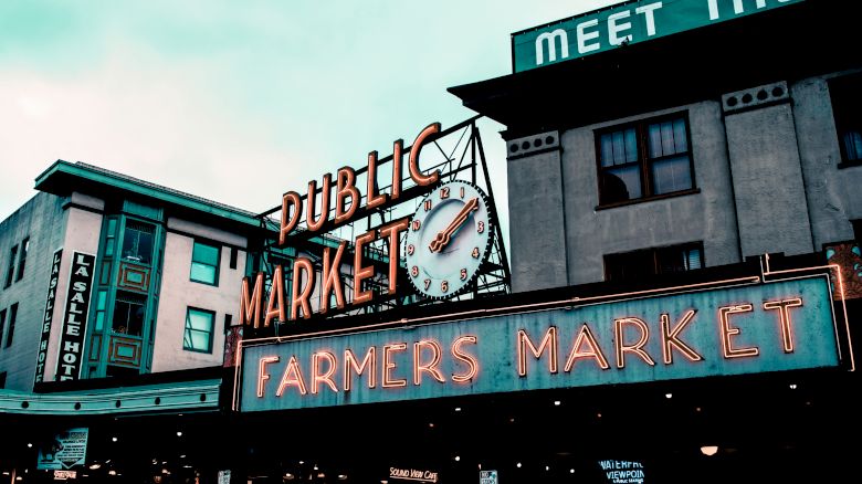 The image shows a public farmers market with neon signs, an iconic clock, and buildings with signs in the background under a cloudy sky.