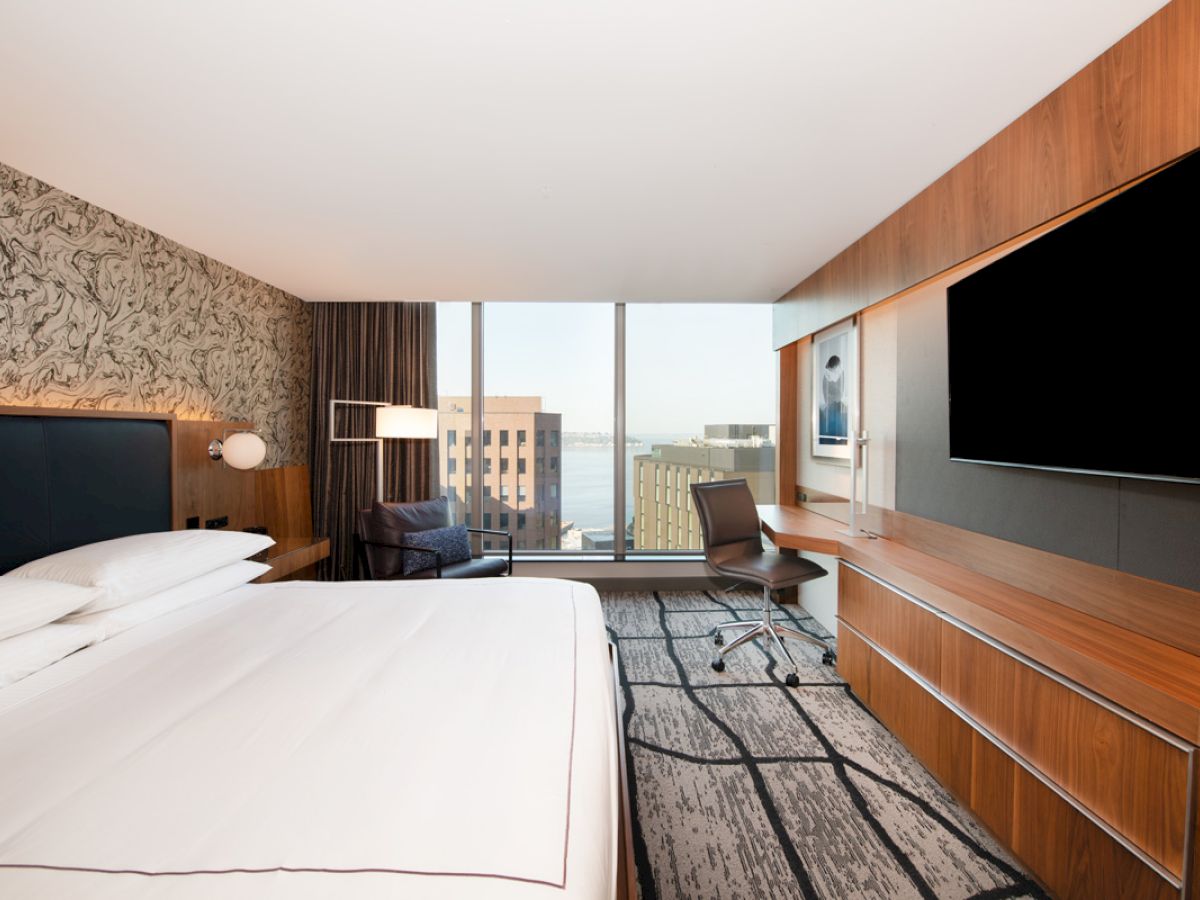 A modern hotel room with a large bed, a desk, a chair, a wall-mounted TV, and a window with a view of a cityscape.