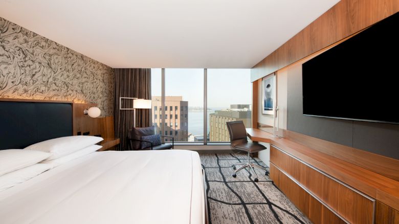 A modern hotel room with a large bed, a desk, a chair, a wall-mounted TV, and a window with a view of a cityscape.