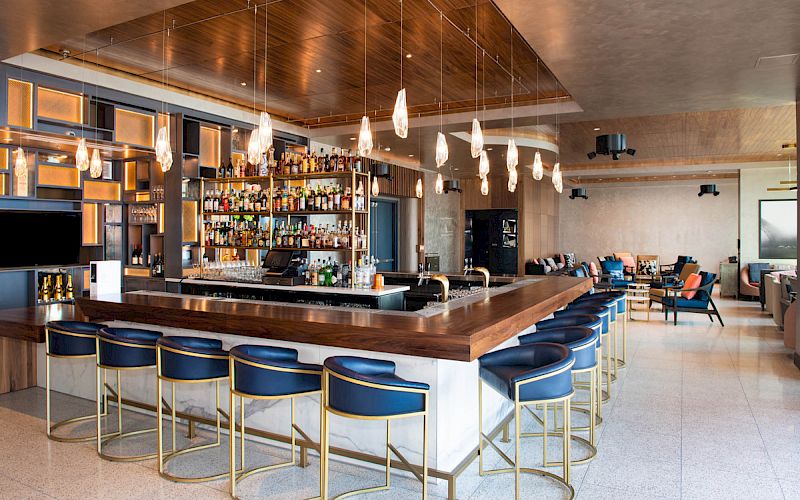 A modern bar with blue stools, a stocked liquor shelf behind the counter, and pendant lights hanging from the ceiling. There are seating areas.