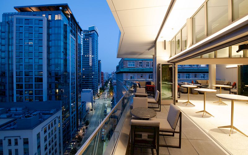 A modern cityscape view from a high-rise balcony at dusk, featuring outdoor seating and large windows, overlooking streets and buildings.