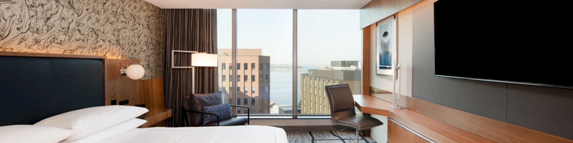 A modern hotel room with a large bed, a desk, a chair, a TV, and a window offering a city view.