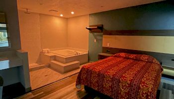 A cozy bedroom with a jacuzzi, bed with a red patterned cover, and striped wooden flooring, lit by soft lighting.