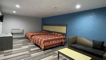 The image shows a modern hotel room with two beds, a couch, a coffee table, and a TV mounted on the wall, featuring a blue accent wall.