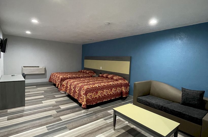 The image shows a modern hotel room with two beds, a couch, a coffee table, and a TV mounted on the wall, featuring a blue accent wall.