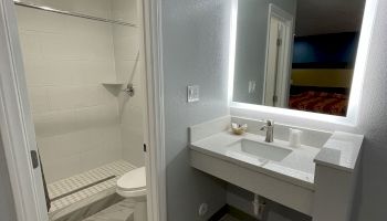 A modern bathroom with a lighted mirror, white countertop sink, and a shower area with a glass door and rain shower head visible.