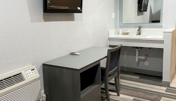 The image shows a room with a wall-mounted TV, a desk with a chair, an air conditioning unit, and a sink with a mirror above it.