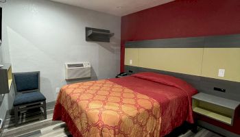 This image shows a tidy hotel room with a double bed, a patterned bedspread, a chair, and a small air conditioning unit on the left wall.