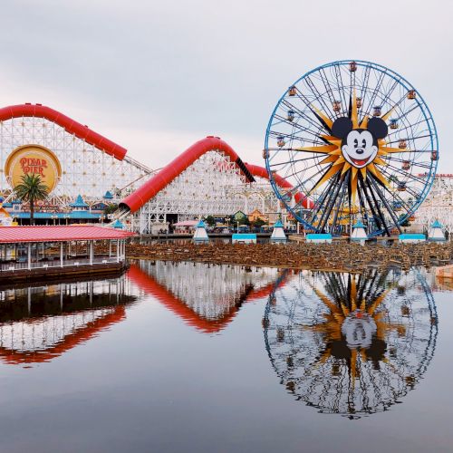 The image shows an amusement park with a large Ferris wheel and roller coaster, reflected in a water body, featuring a prominent animated character.