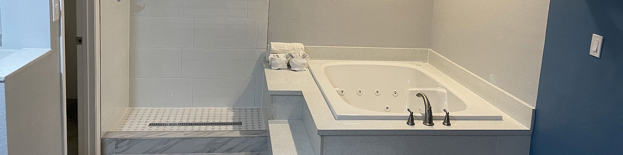 The image shows a modern bathroom with a bathtub, walk-in shower, and white tile flooring. The room features blue walls and recessed lighting.